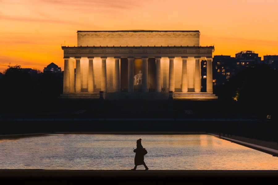 La silhouette d'une personne marchant le long du bassin réfléchissant se détache sur le Lincoln Memorial illuminé au coucher du soleil, avec un ciel orange chaud et des bâtiments de la ville en arrière-plan.