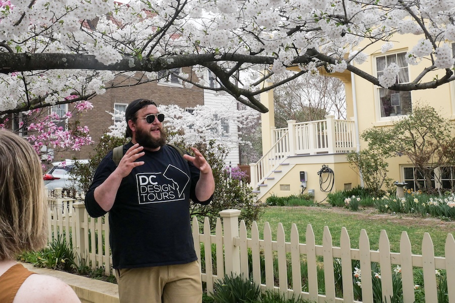 A tour guide speaks outside of a historic home with a blooming cherry blossom tree behind him
