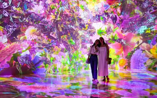 Two visitors stand in a vibrant, immersive digital art installation at ARTECHOUSE, surrounded by colorful, glowing flowers and butterflies.

