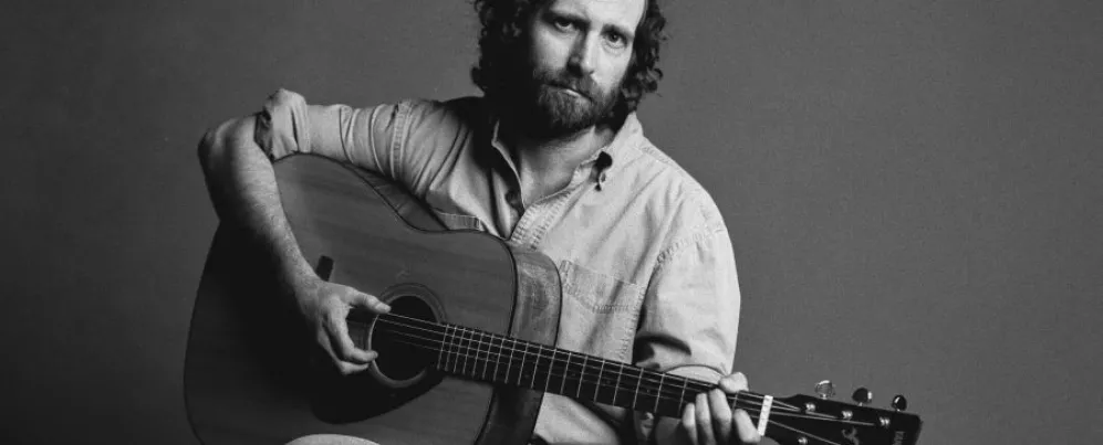 Comedian and musician Kyle Mooney poses with an acoustic guitar in a black and white promotional portrait.